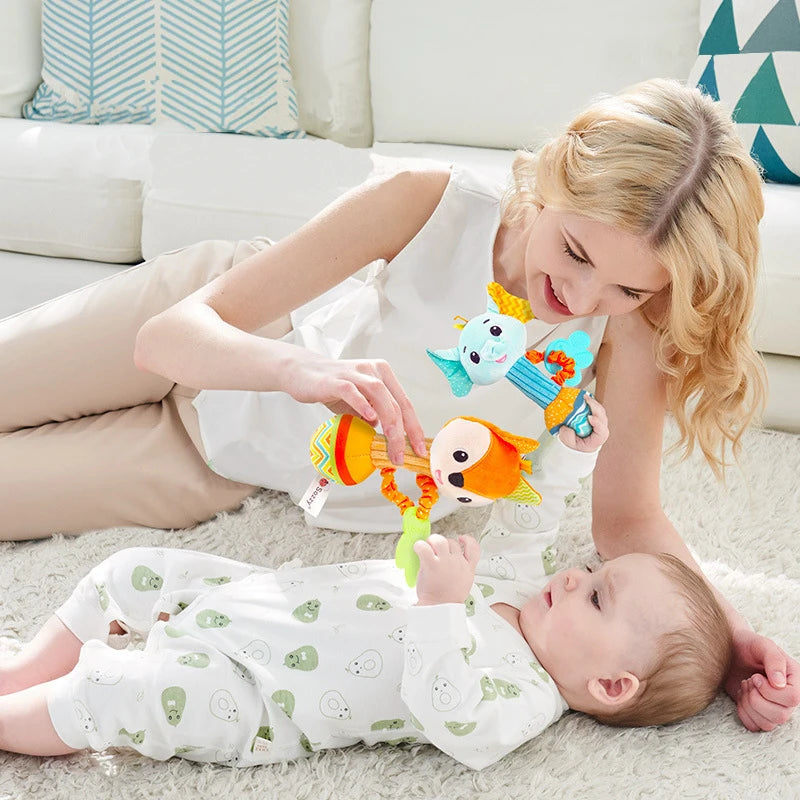 une femme aux cheveux long blonds ondulés, en chemisier sans manche blanc et un pantalon beige et un bébé en pyjamas blanc avec motifs avocat blanc et vert allongés jouant avec deux hochets éléphant et renard sur un tissu à poils long clair avec des coussins bleus et blanc derrière