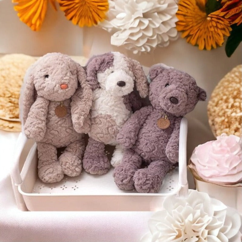 peluche lapin beige avec pendentif marron, peluche chien tricolore blanc beige et marron à coté d'un ours marron avec un pendentif marron dans un support blanc