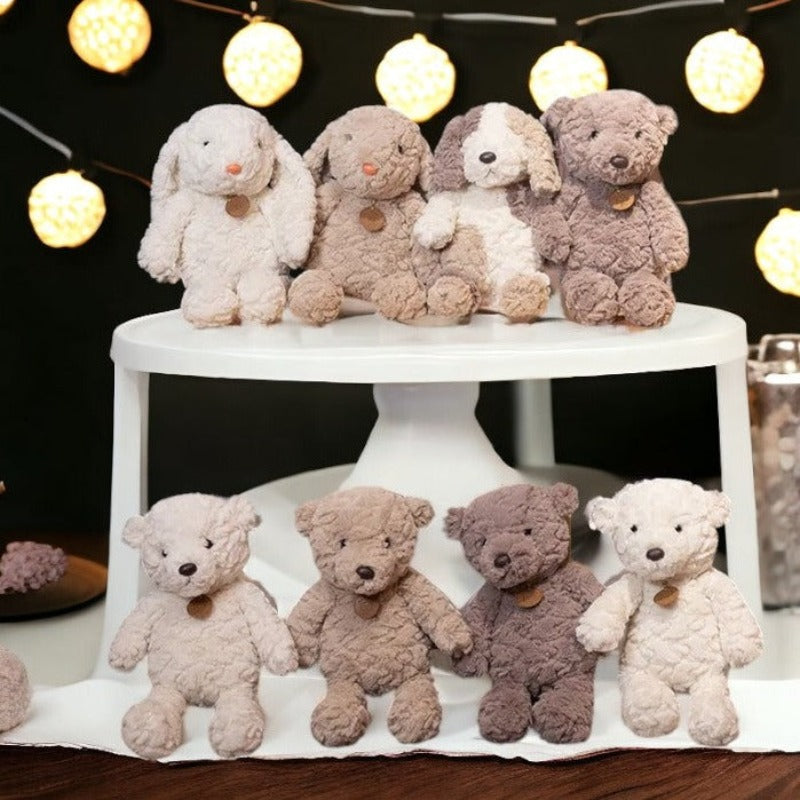 peluche lapin blanc et peluche lapin  beige avec pendentif marron, peluche chien tricolore blanc beige et marron à coté d'un ours marron avec un pendentif marron sur une table blanche et quatre ours beige, crème, marron et blanc , tous avec pendentif. Posés sur tissu blanc 