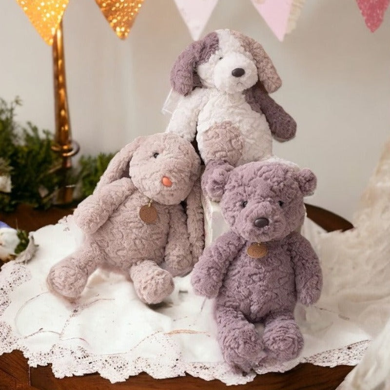 peluche lapin beige avec pendentif marron, peluche chien tricolore blanc beige et marron à coté d'un ours marron avec un pendentif marron posé sur un tissu blanc sur une table en bois