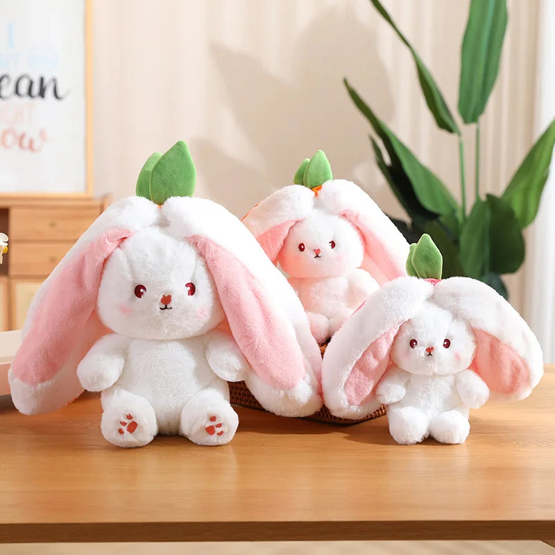 trois peluches lapins blancs de trois dimensions différente posé en triange sur une table en bois dons un posé sur un panier en rotin, derrière à gauche un tableau posé sur deux tiroirs en bois et une plante verte à gauche