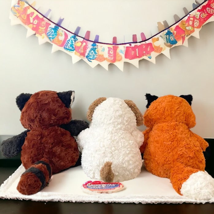 peluche raton laveur marron blanc et noir, peluche chien blanc et marron aux cotés de la peluche renard orange , blanche et noir ( de dos )