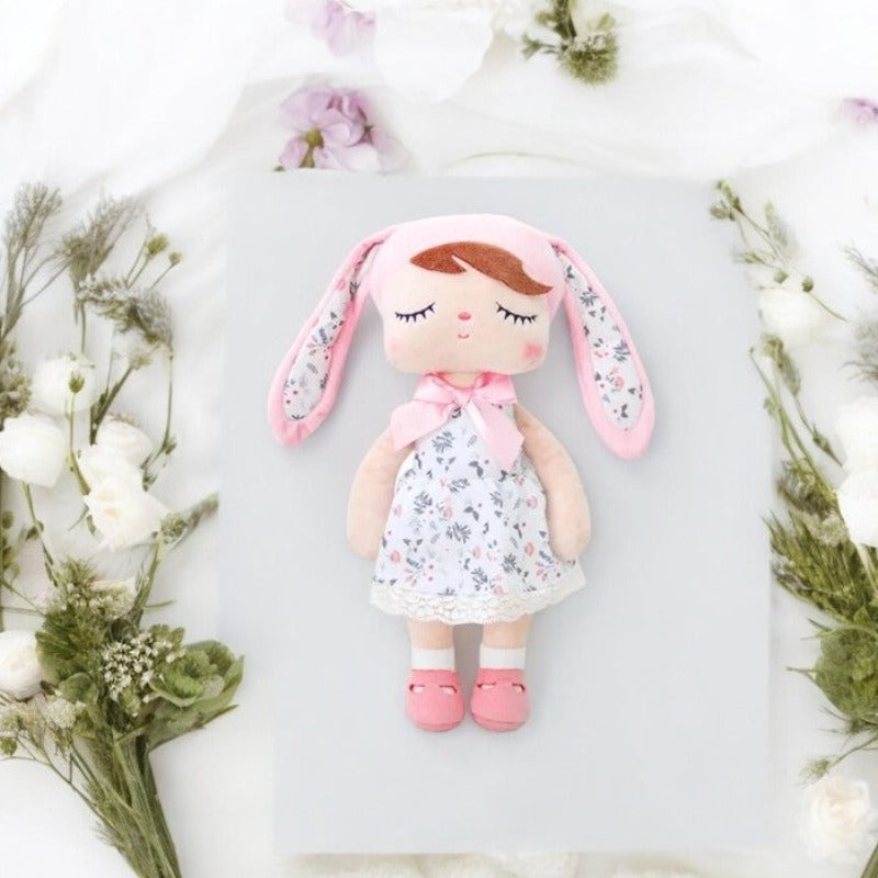 SURI, la fée dolls de 34 cm, éblouit avec sa robe blanche à fleurs, son bonnet à oreilles de lapin rose et blanc assorti, et ses ballerines roses, créant ainsi un tableau enchanté digne des contes de fées.