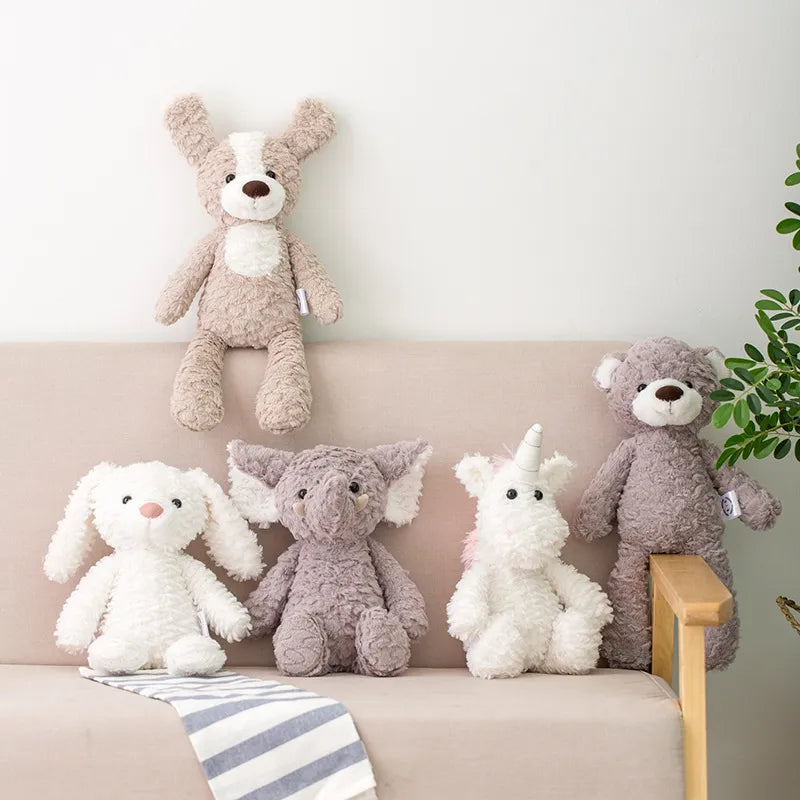 peluche chien bicolore posée sur le dossier du canapé beige, lapin blanc posé sur une bande de tissu rayée bleu et blanc sur le canapé, l’éléphant gris, suivie de la licorne blanche et sur l’accoudoir en bois un ours gris est installé à califourchon