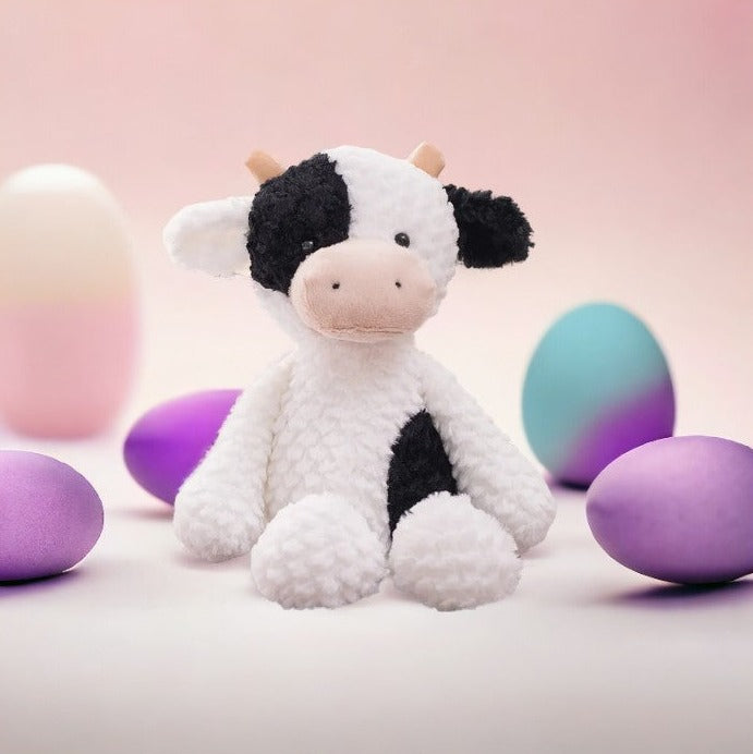 peluche vache blanche et noire assisi avec un décors dans les tons roses avec des formes œufs rose bleu et violet derrières elle