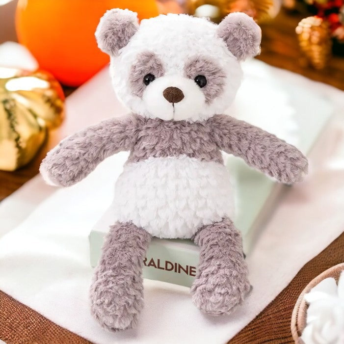 peluche panda grise et blanche posée sur un livre blanc, sur un tissu blanc avec des objets or et orange derrière 