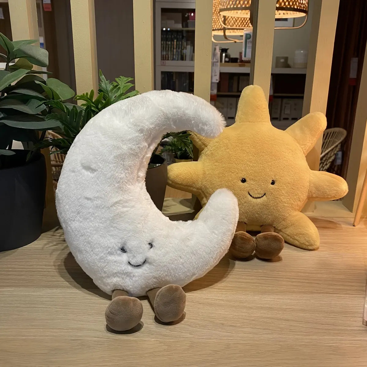 peluche lune souriante blanche avec peluche soleil jaune souriant, sur table en bois avec plantes vertes