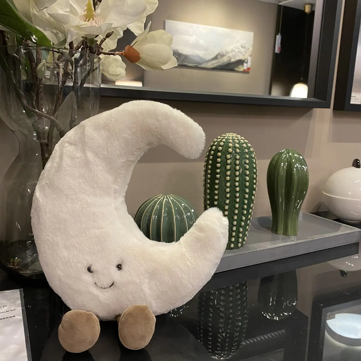 peluche lune souriante blanche sur table en verre avec cactus artificiels au revêtement brillant