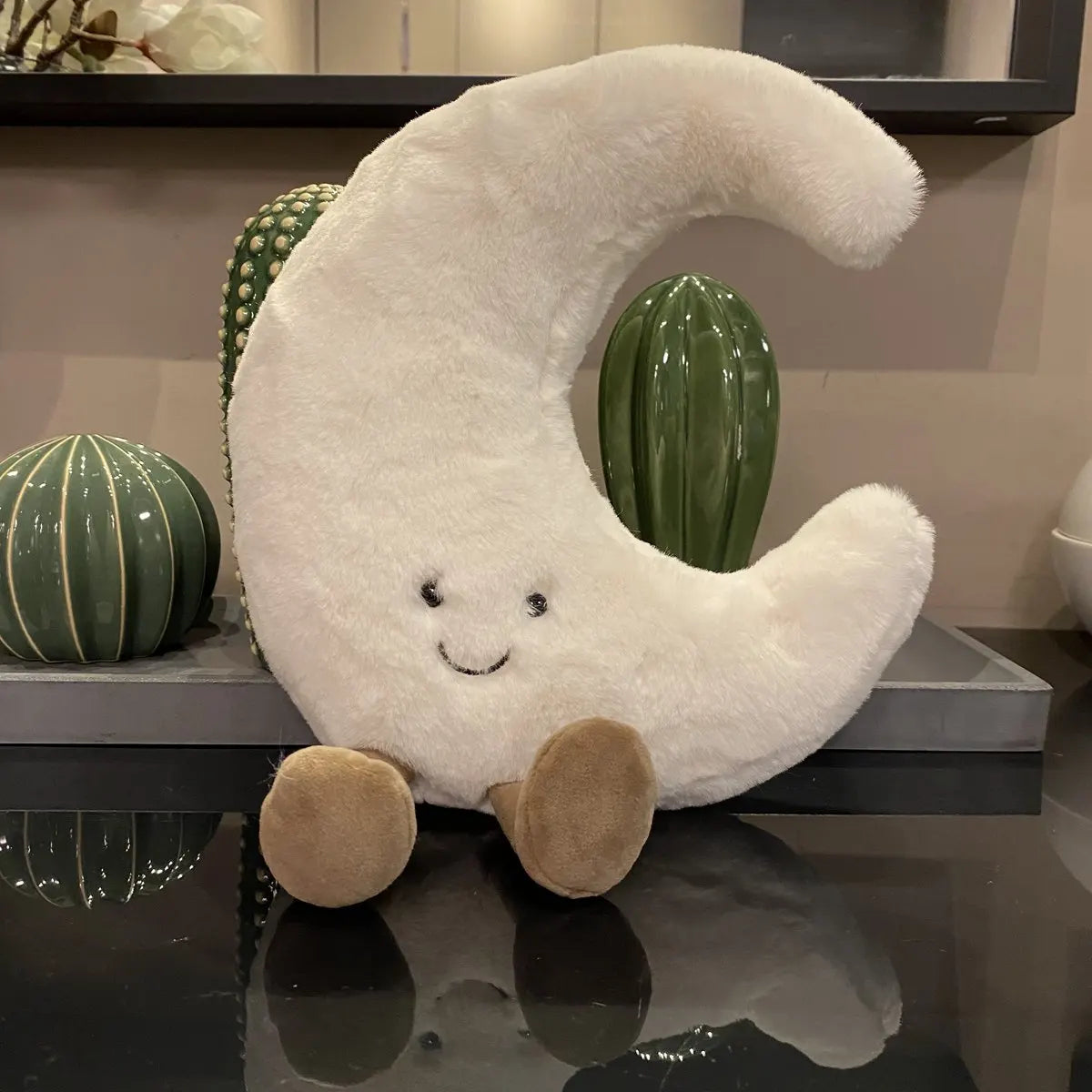 peluche lune blanche posée sur une table ne verre devant un plateau de petit cactus en résine brillante.