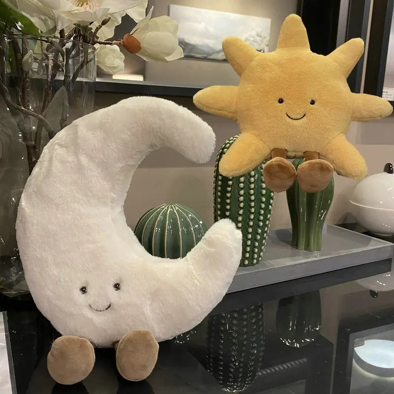 peluche lune souriante blanche avec peluche soleil jaune souriant, sur table en verre avec cactus artificiels au revêtement brillant