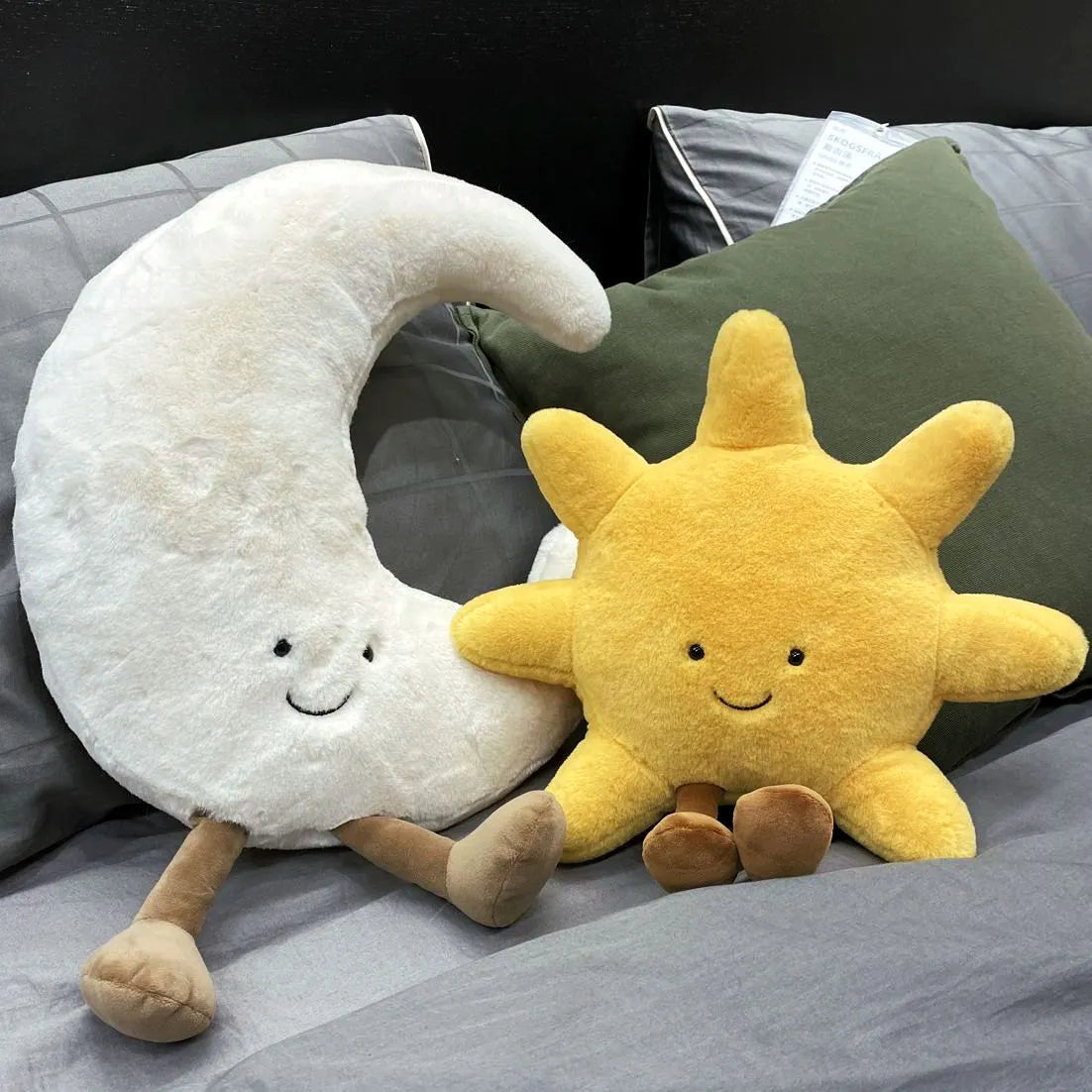 peluche lune souriante blanche avec peluche soleil jaune souriant, posées parmi des  coussins gris bleutés